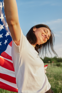 A person smiling outdoors, holding an American flag draped over their shoulders, with a blue sky and green field in the background.
