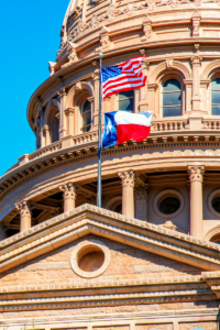 The American and Texas state flags flying in front of the dome of the Texas State Capitol building on a clear day.