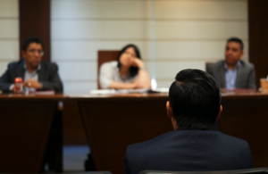 A person faces three people seated at a table in a meeting or interview setting, with papers and drinks on the table.
