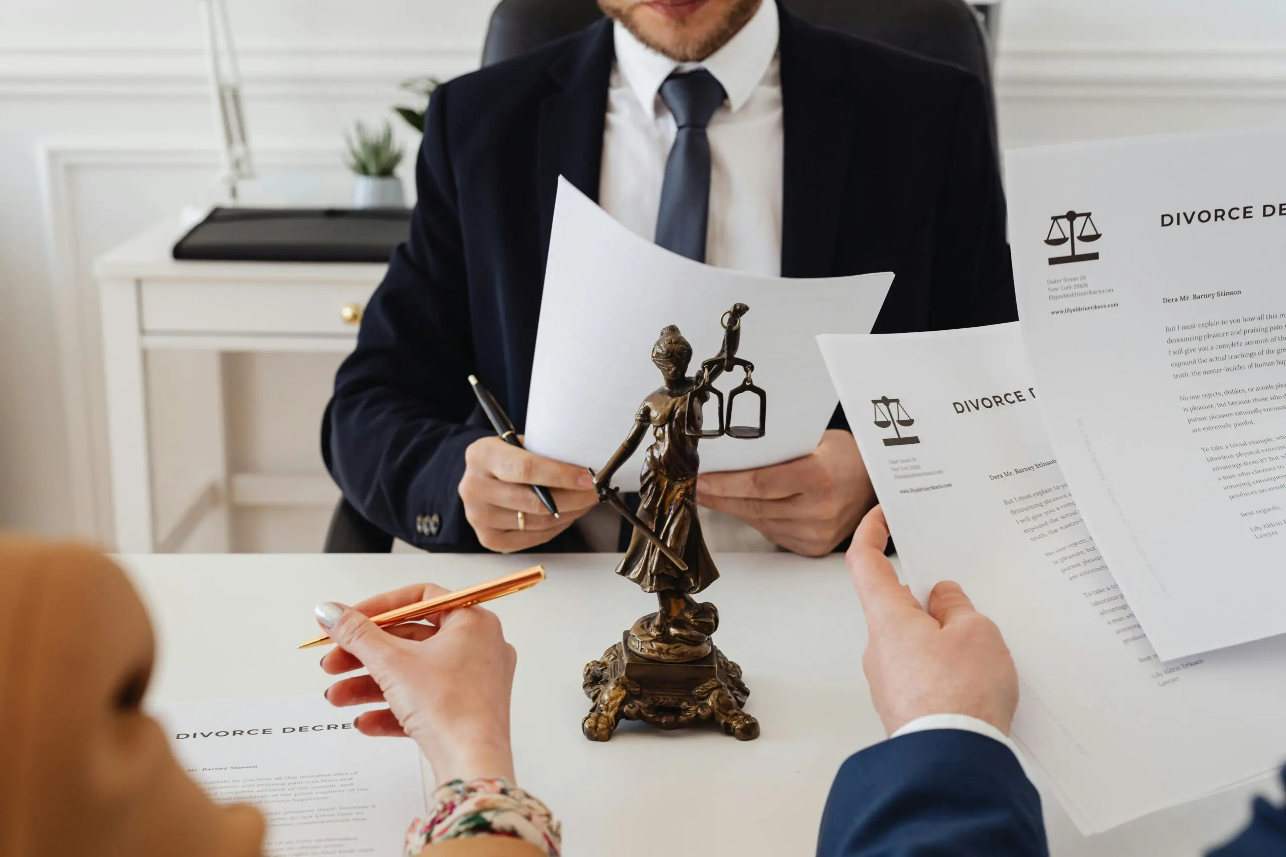 Three people sit at a desk with divorce documents and a Lady Justice statue, discussing legal paperwork in a professional office setting.