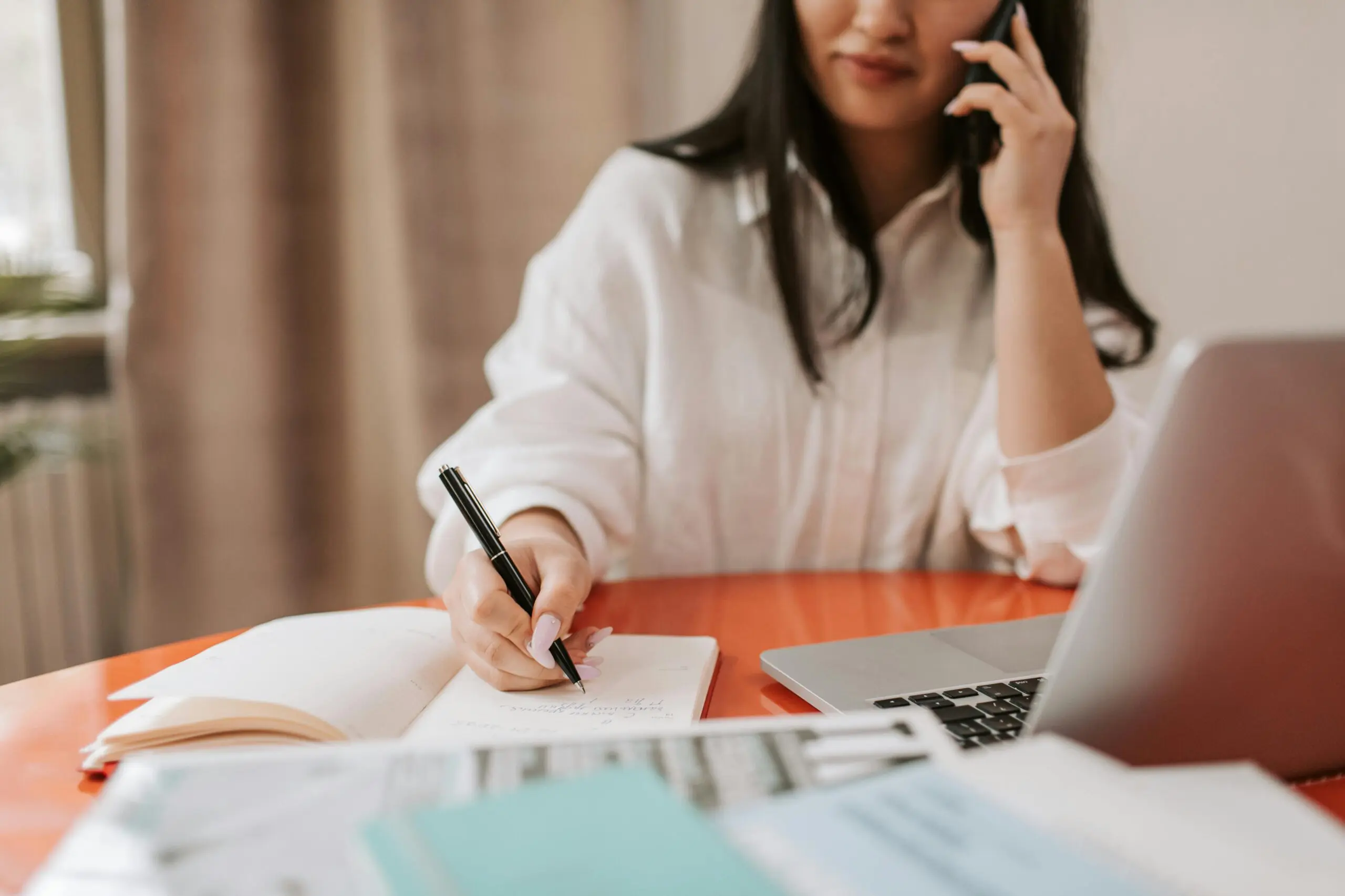 Woman talking on the phone, writing in a notebook, and working at a laptop on a table with papers.