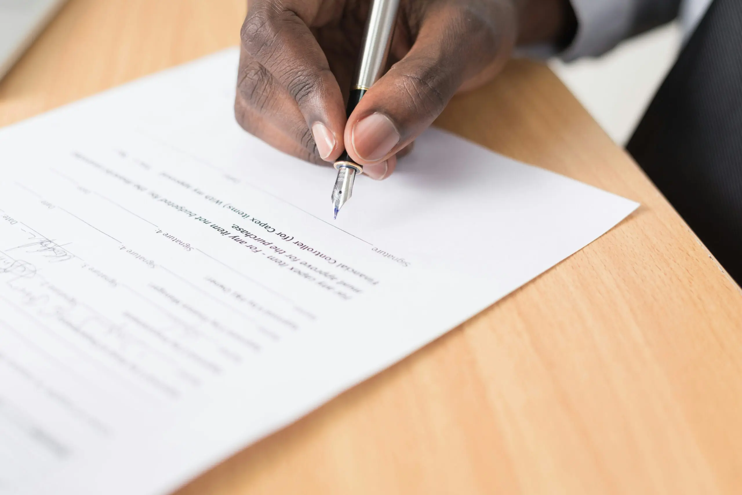 A person’s hand holding a pen and signing a printed document on a wooden desk.