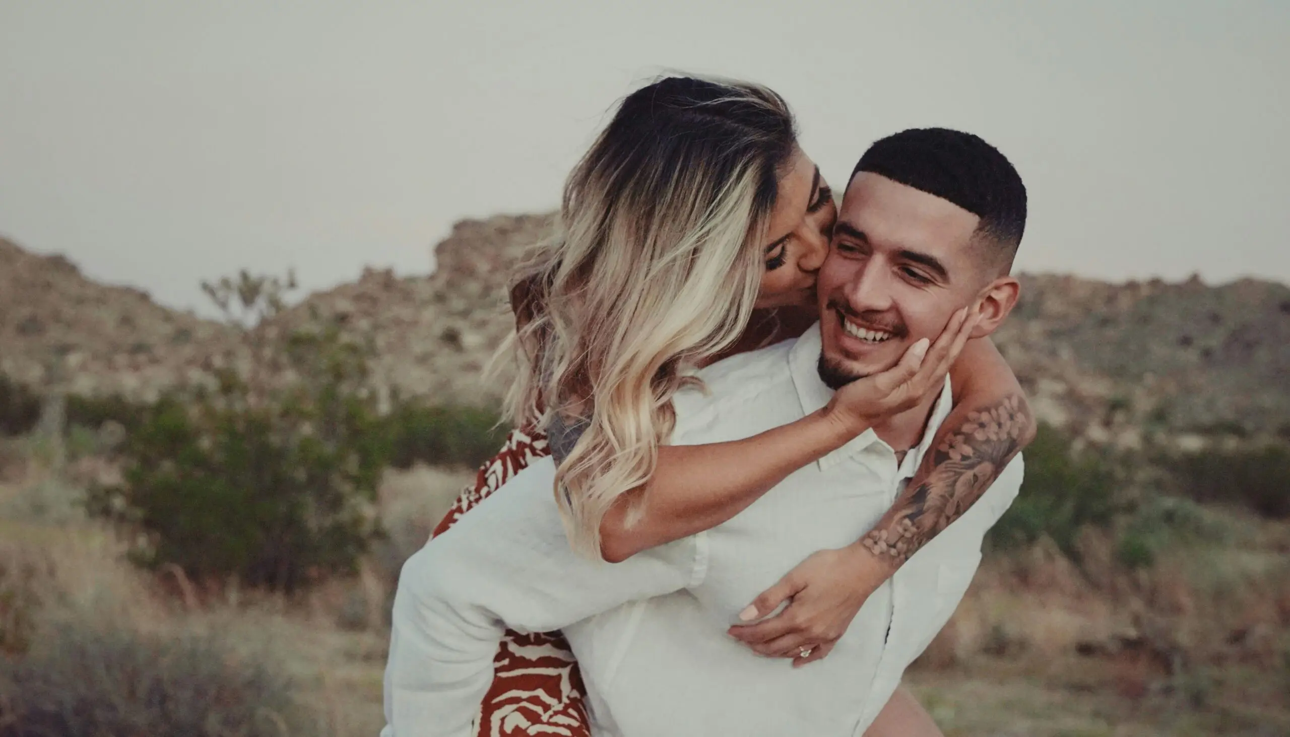 A woman kisses a smiling man on the cheek while giving him a piggyback ride outdoors with rocky hills and vegetation in the background.