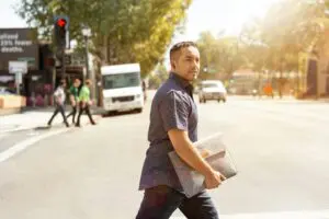 Man crossing a city street in daylight holding a laptop sleeve, with cars and pedestrians visible in the background.