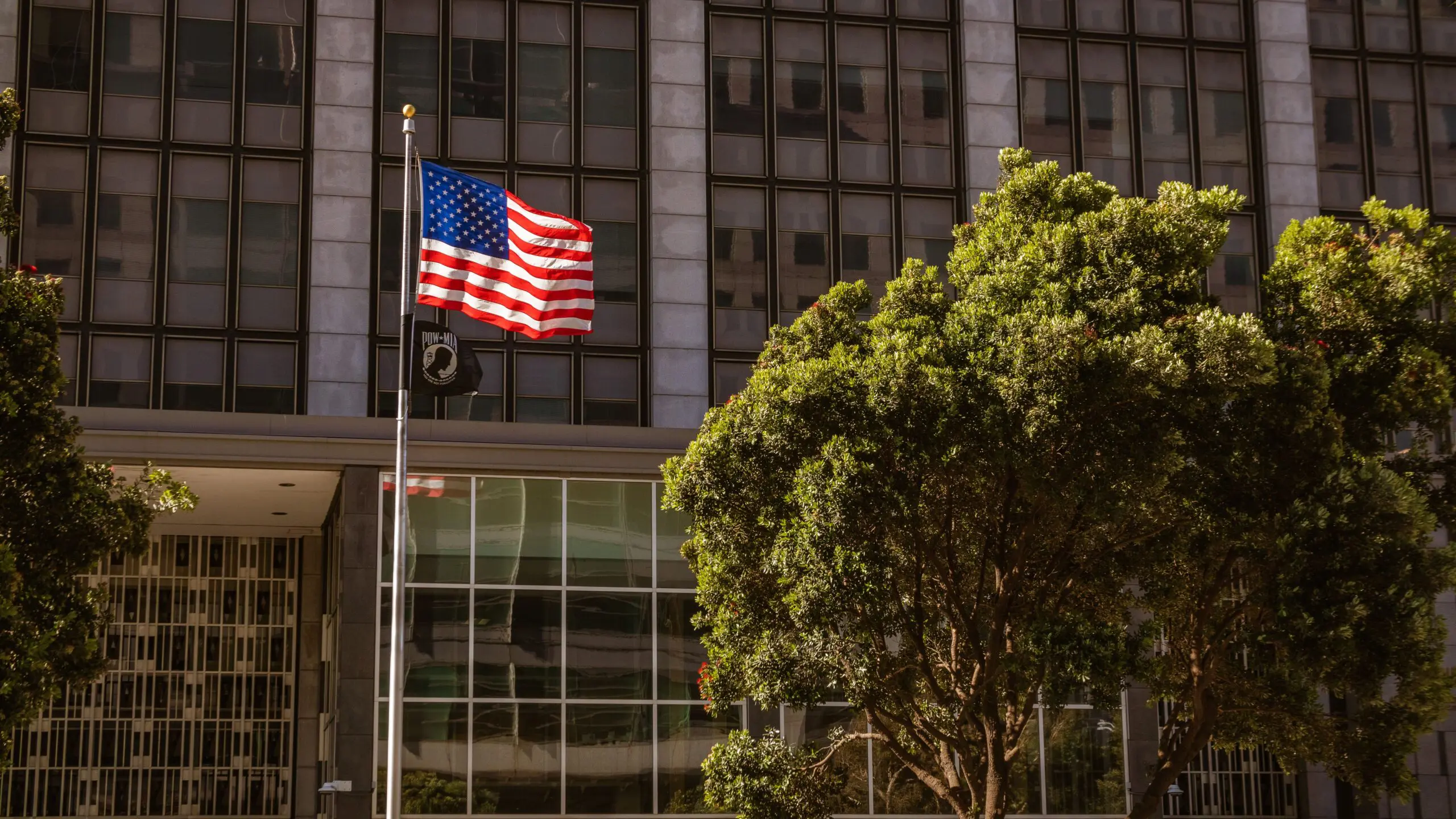 An American flag and a POW/MIA flag flying in front of a modern office building with large windows and trees in the foreground.