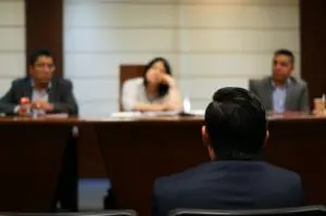 A person sits facing three people at a conference table in a formal meeting room. The three individuals appear to be listening or evaluating.