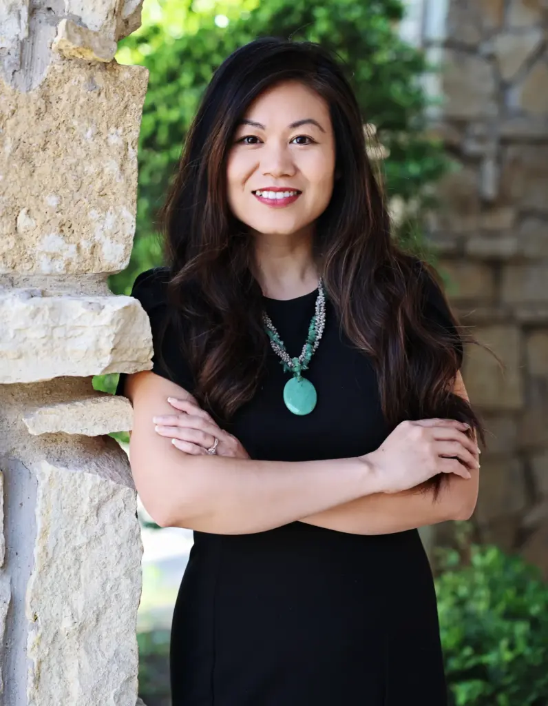 Woman with long dark hair in a black dress stands with arms crossed, smiling, in front of a stone wall and greenery.