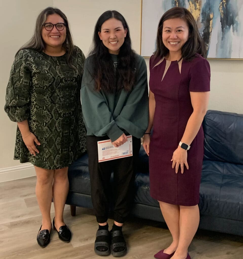 Three women stand indoors, smiling at the camera; the woman in the center holds an envelope while the other two stand on either side of her.