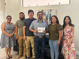Six people stand together indoors, smiling at the camera; the man in the center holds a document and envelope. A couch and artwork are visible in the background.