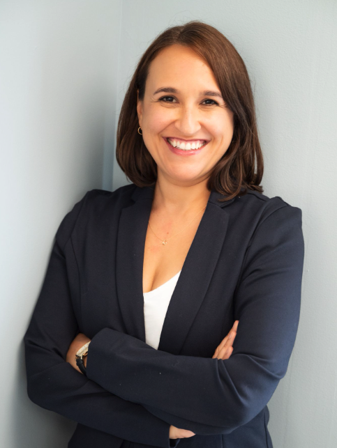 Woman in a dark blazer and white top stands against a light-colored wall with arms crossed, smiling at the camera.