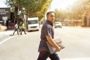 Man crossing a city street in daylight holding a laptop sleeve, with cars and pedestrians visible in the background.