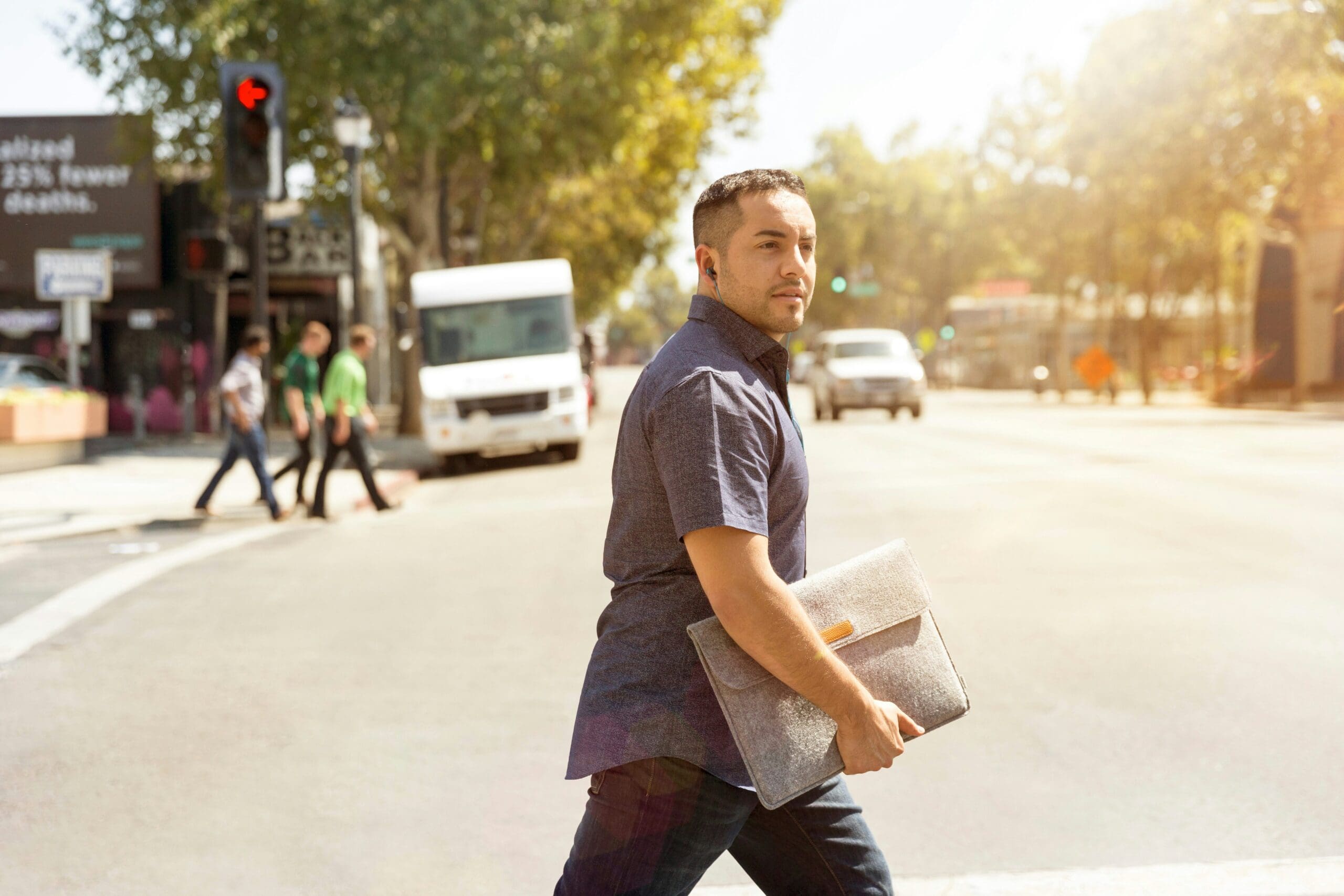 Man crossing a city street in daylight holding a laptop sleeve, with cars and pedestrians visible in the background.