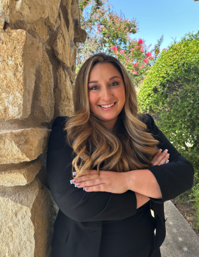Woman with long, wavy hair and a black outfit stands with arms crossed by a stone wall, outdoors on a sunny day with greenery and flowering shrubs in the background.
