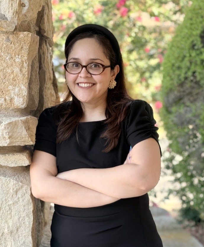 Woman with glasses and dark hair, wearing a black dress and headband, stands with arms crossed by a stone wall outdoors, with greenery and flowers in the background.