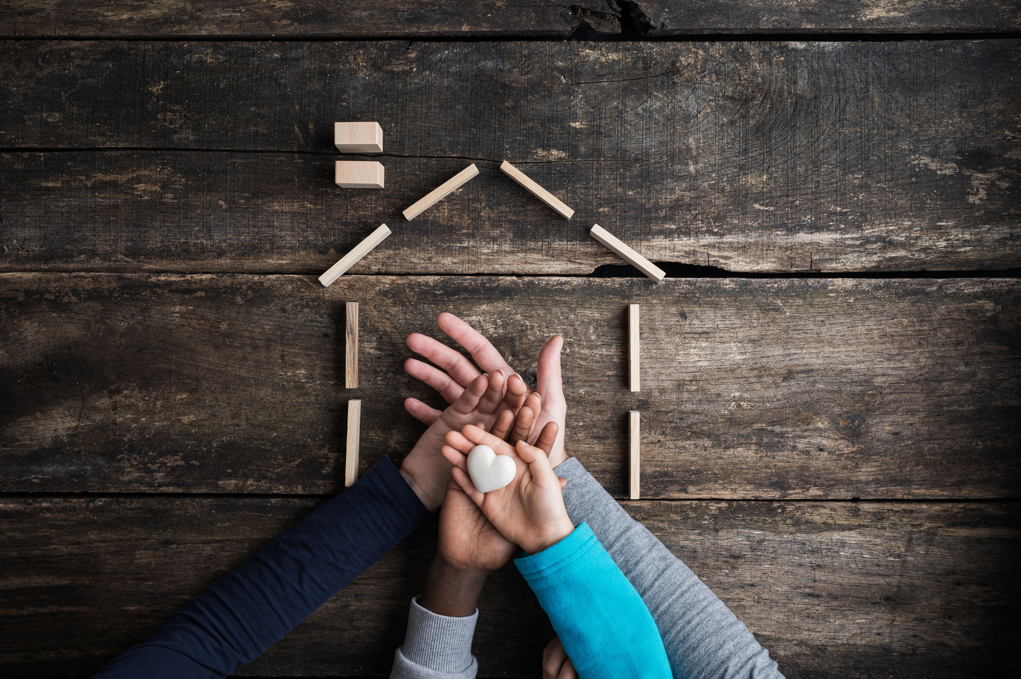 Several hands of different skin tones stack together holding a small heart inside a house-shaped outline, representing the family safety and reunification a T visa can offer trafficking survivors.